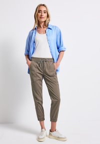 Chemise boutonnée bleu clair sur un débardeur blanc, associée à un pantalon fuselé vert olive. La tenue comprend des poches et une taille élastique avec cordon.
