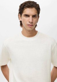 Ung mand med mellemlangt brunt hår, iført en enkel hvid T-shirt, står imod en lys baggrund med en neutral udtryk.