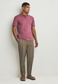 Ružové polo tričko s krátkymi rukávmi a jemným logom, v kombinácii s béžovými nohavicami na mieru a tmavozelenými sandálmi. Minimalistický dizajn.