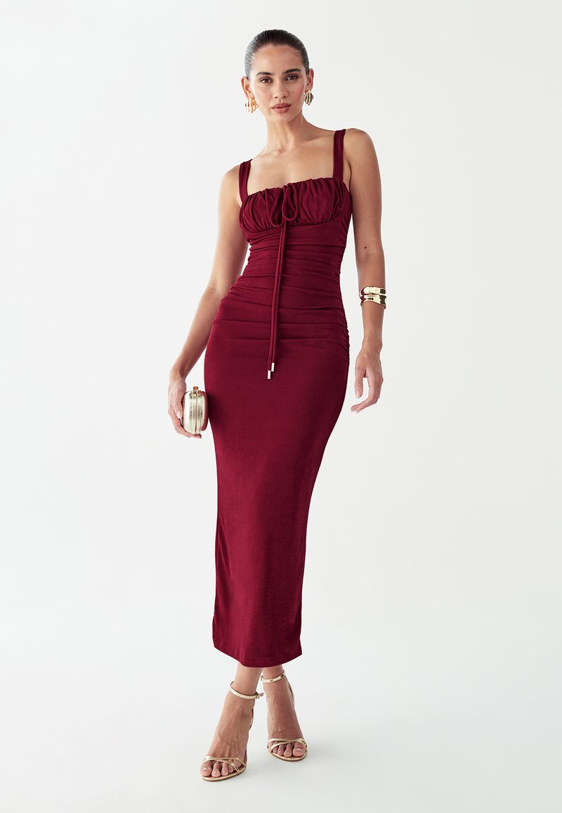 Vestido bodycon burdeos con fruncidos, escote cuadrado y tirantes finos. Combina con accesorios dorados y zapatos de tacón con tiras, acompañado de un pequeño bolso de mano.