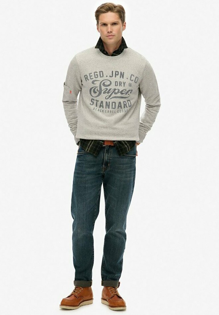 Hombre de pie con las manos en los bolsillos, vistiendo un suéter gris con texto, una camisa de cuadros oscuros, jeans enrollados y botas de cuero marrón.