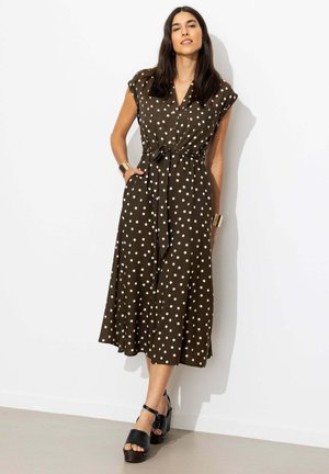 Femme debout contre un mur blanc, portant une robe midi marron à pois blancs, taille cintrée avec ceinture, manches courtes, talons compensés noirs et bracelets dorés.