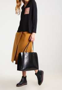 Borsa tote in pelle nera con manici lunghi, texture liscia e due chiusure superiori; indossata con una camicia nera oversize e una gonna senape.