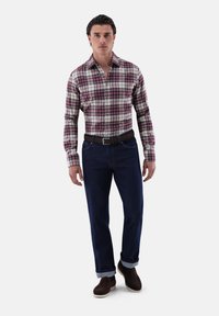 Camicia a quadri in rosso, blu navale e bianco; chiusura frontale a bottoni. Jeans blu scuro con risvolto. Scarpe marroni senza lacci. Cintura semplice marrone.