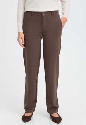 Femme portant un pantalon marron droit, un pull crème et des chaussures plates marron foncé, debout avec une main dans la poche devant un fond uni.