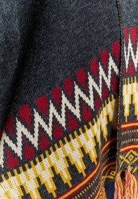 Pleta pončo s námorníckou základnou, zdobené výrazným geometrickým vzorom v červenej, žltej a krémovej farbe. Dekoratívne třásně na leme.