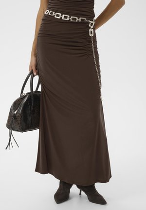 Femme portant une jupe froncée marron avec une ceinture chaîne argentée, tenant un sac à main noir texturé et portant des bottines noires à talons pointus.