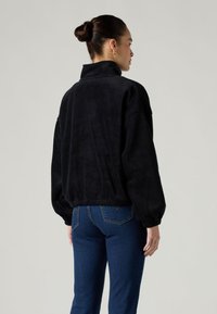 Μαύρο πουλόβερ από φλίς με ψηλό γιακά, κατεβασμένους ώμους και ελαστικές μανσέτες. Φορεμένο με στενά μπλε τζιν, που δείχνουν μια άνετη εφαρμογή.