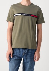 Muž v olivově zeleném tričku Tommy Hilfiger s logem napříč hrudí, doplněný modrými džíny, stojící na bílém pozadí.