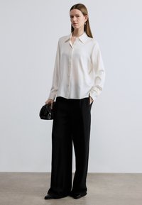 Long-sleeve white silk shirt with button-down front, paired with loose black trousers, soft black handbag, and pointed black shoes.