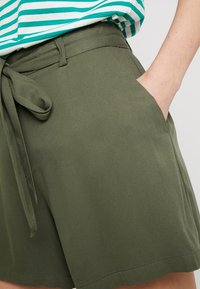 Pantaloni corti verde oliva con vita alta, cintura in tessuto e tasche laterali. La texture appare liscia con una vestibilità su misura.