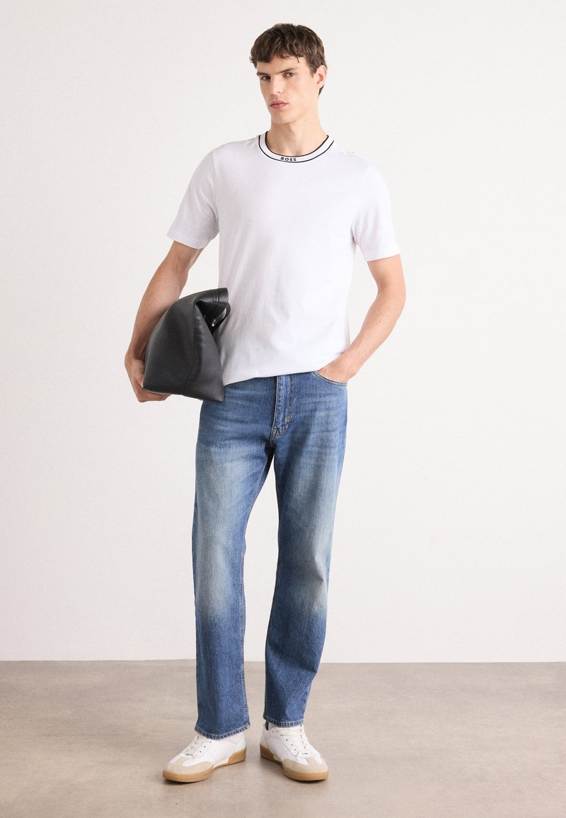 Weißes T-Shirt mit kurzen Ärmeln und schwarzem Kragenakzent, hellblaue Jeans und weiße Sneakers mit beigen Sohlen; hält eine schwarze Tasche.