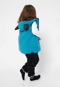 Teal-Fleece-Weste mit Kapuze, mit schwarzen Akzenten und Ohren. Getragen über einem weißen Langarmshirt und schwarzen Plüschhosen.