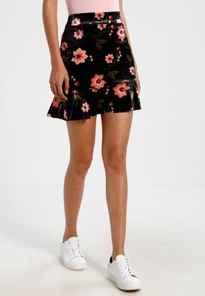 Zwarte bloemenrok met een gerafelde zoom, met roze en rode bloemen en groene bladeren, gemaakt van een gestructureerde stof. Draagt witte sneakers.