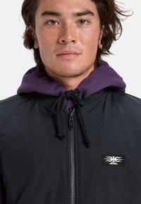 Giacca nera con cappuccio, foderata in viola, caratterizzata da una zip frontale e un patch con il logo. Texture liscia e cappuccio con cordino regolabile.