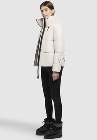 Veste blanche rembourrée avec col haut et fermeture éclair noire, portée sur des leggings noirs et des bottines noires à lacets, mettant en avant un design épuré.