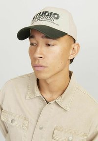 Beige kasket med en mørkegrøn skygge, med teksten "STUDIO Originals" i sort og hvid. Båret med en beige skjortejakke.