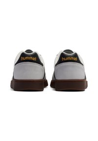 Un paio di scarpe da ginnastica bianche e grigie con suole marroni, viste da dietro, con pannelli del tallone neri e il logo "hummel" in oro.