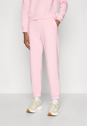 Pink sweatpants with elastic cuffs, made of soft fabric. Features a subtle logo on the front and a relaxed fit. Worn with white sneakers.