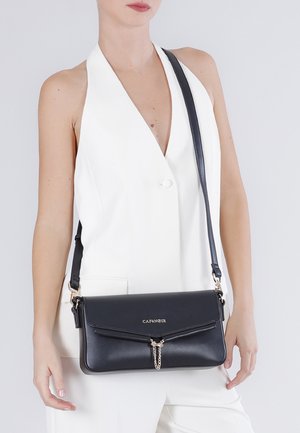 Kvinde iført en hvid ærmeløs blazer og bærende på en sort læder crossbody-taske med kædedetalje og "CAFENOIR"-logo.