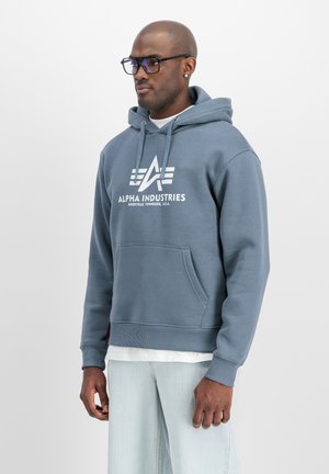 Mann trägt blauen Alpha Industries Hoodie mit Fronttasche, helle Jeans, Brille und Ohrringe, steht vor einem schlichten hellen Hintergrund.