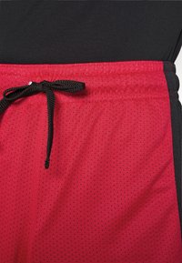 Primer plano de pantalones cortos deportivos rojos con perforaciones y cordón negro atado en la cintura, llevados sobre leggings negros.