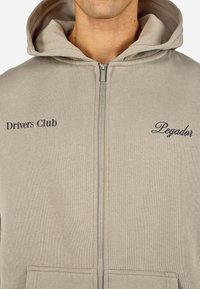 Felpa zip con cappuccio beige, dotata di tasca frontale. Presenta le scritte "Drivers Club" e "Pagador" stampate in caratteri scuri sul petto. Texture morbida.