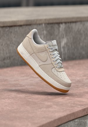 Baskets Nike Air Force 1 beiges et blanches avec semelle en caoutchouc flottant au-dessus d'une surface en pierre rose et d'un fond en pierre grise.