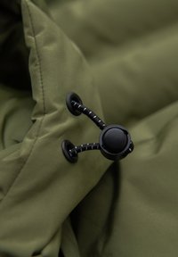 Fermeture à bascule en plastique noir fixant un cordon réglable sur un tissu matelassé vert, probablement partie d'une veste ou d'un manteau.