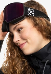 Nuori nainen, jolla on kiharat hiukset, säätää mustia Roxy-lyhtylaseja, päällään musta takki valkoista taustaa vasten.