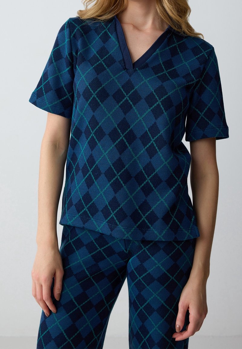 Femme portant un haut à manches courtes bleu et vert à motif losanges et un pantalon assorti, debout devant un fond clair uni.