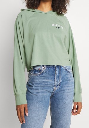 Kobieta ubrana w jasnozieloną, krótką bluzę z kapturem z logo "Hollister" oraz wysokowysokie niebieskie dżinsy, stojąca na tle jednolitego tła.