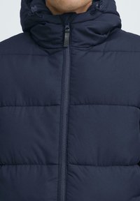 Marineblå pufferjakke med høy krage, glidelås foran og kviltet tekstur. Har justerbare snorer på hetten.