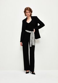 Blazer nero su una maglietta nera e pantaloni a gamba larga, con una cintura bianca a motivi e tacchi alti neri. Tessuto dalla consistenza liscia.