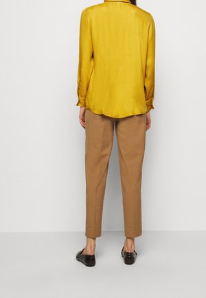 Persona in piedi di spalle che indossa una camicia a maniche lunghe giallo senape, pantaloni dritti color cammello e mocassini neri su uno sfondo semplice.