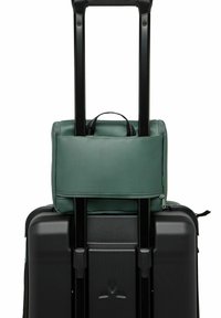 Zwarte harde koffer met een textuur op het oppervlak, bedekt met een groene stoffen tas met een handvat, die om in- en uitschuifbare zwarte handvatten is bevestigd.