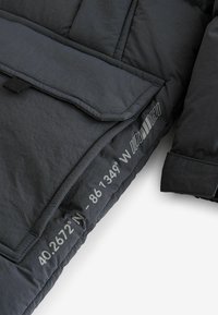 Black puffer jacket with a textured surface, featuring a large flap pocket and printed coordinates in light grey on the side.
