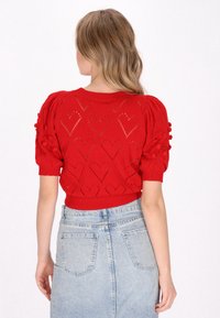 Rød strikket sweater med pufærmedesign, hjerteformede udskæringer og teksturerede pom-pom detaljer; parret med en lys blå denimnederdel.