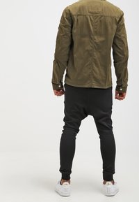 Olive green jacket with a buttoned front and a pocket on the left shoulder, paired with black joggers and white sneakers.