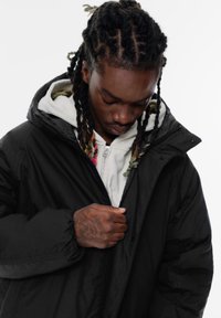 Cappotto isolato nero con cappuccio, caratterizzato da una chiusura frontale con zip e polsini elasticizzati, indossato sopra una felpa bianca con fodera multicolore.