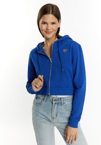Felpa blu a zip corta in morbido tessuto con un logo a forma di cuore sul petto, abbinata a jeans a vita alta di un lavaggio chiaro.