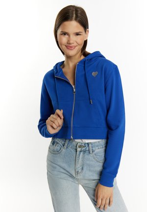 Geknipte blauwe zip-up hoodie van zachte stof met een hartlogo op de borst, gecombineerd met high-waisted lichtgekleurde spijkerbroeken.