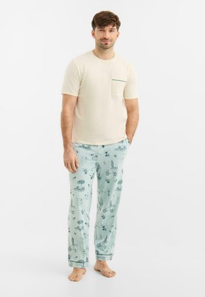 Man die een beige korte mouwen shirt en lichtblauwe pyjamabroek draagt met donkerblauwe zomerse illustraties, stond blootsvoets op een witte achtergrond.