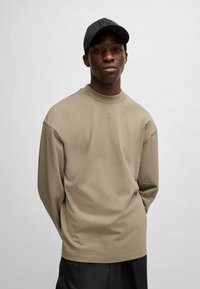 Beige Langarmhemd mit rundem Ausschnitt, ausgestattet mit dezenten Logodetails in heller Stickerei, kombiniert mit einer schwarzen Mütze. Glatte Textur.