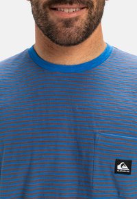 Pruhované tričko s modro-šedými horizontálními pruhy, kulatým modrým výstřihem a kapsou na levé části hrudi s logem. Bavlněný materiál.