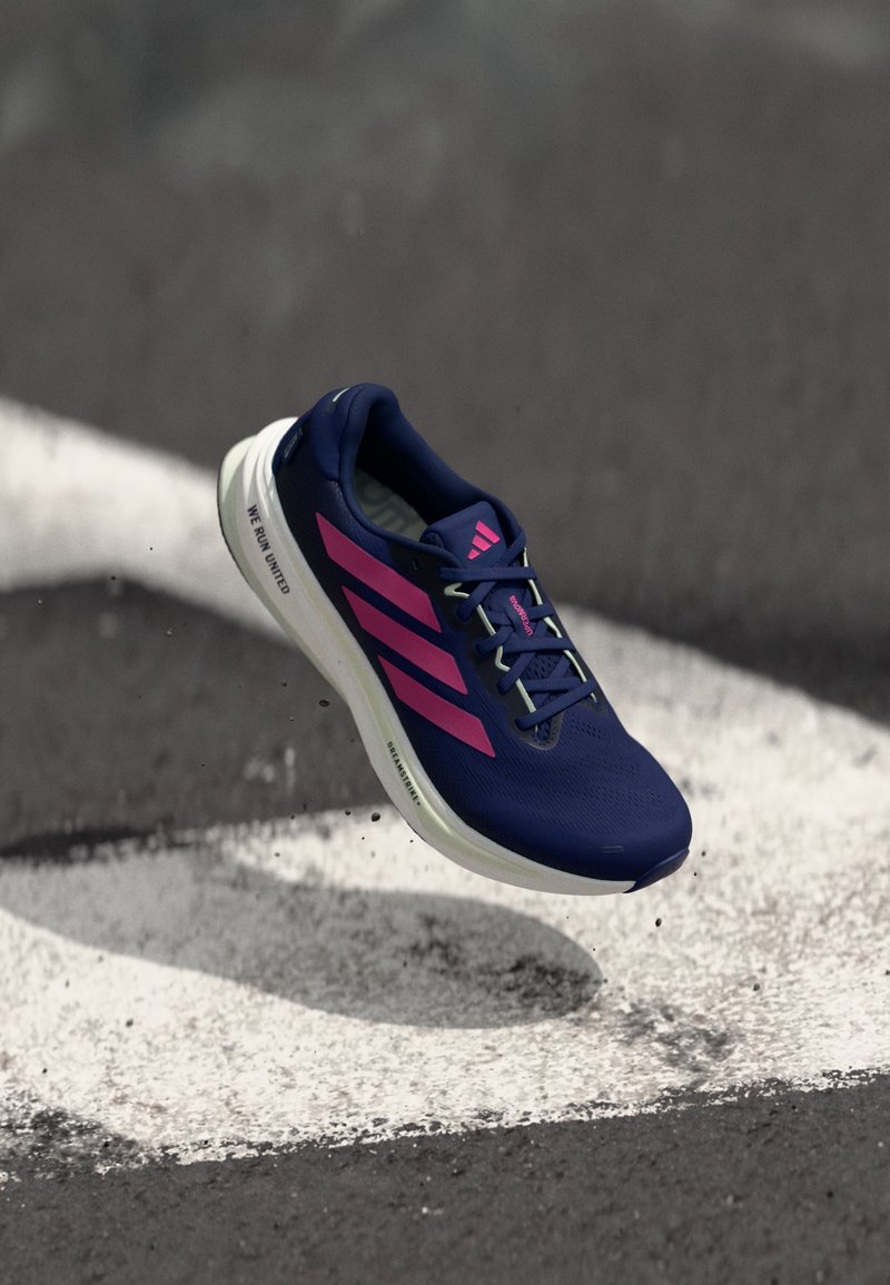 Chaussure de course Adidas bleu marine avec des bandes roses, capturée en plein air au-dessus de l’asphalte avec un marquage au sol blanc et une légère ombre en dessous.