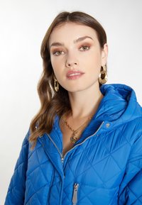 Jachetă albastră cu quilt, cu glugă, cu fermoar în față, detalii argintii și model în diamante. Cercei rotunzi din aur și coliere suprapuse vizibile.