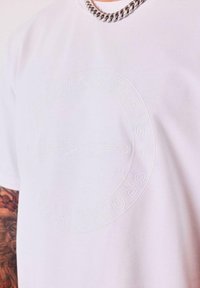 Camiseta blanca con el logo circular en relieve "PROJECT PARIS" y las letras "PXP" en el centro, llevada por una persona tatuada que lleva un collar de cadena de plata.