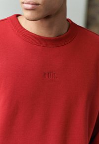 Rød sweatshirt med ribbet halsudskæring og præget "EDIT." logo. Stoffet har en glat, blød tekstur, der er velegnet til hverdagstøj.