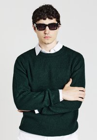 Giovane uomo con capelli ricci e scuri che indossa occhiali da sole neri rettangolari, un maglione a maglia verde scuro sopra una camicia bianca a colletto, in posa con le braccia incrociate.